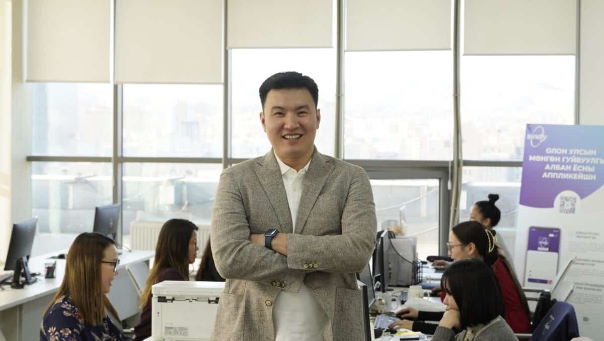 Энтрепренер том цонхтой оффисдоо гараа зөрүүлэн инээмсэглэн өөртөө итгэлтэй зогсож байна. Түүний ард хэд хэдэн хамт олон компьютер дээрээ ажиллаж байгаа харагдана. Оффис нь гэрэлтүүлэг сайтай, орчин үеийн, цонхоор хотын өнгө үзэмжтэй. - Business.MN
