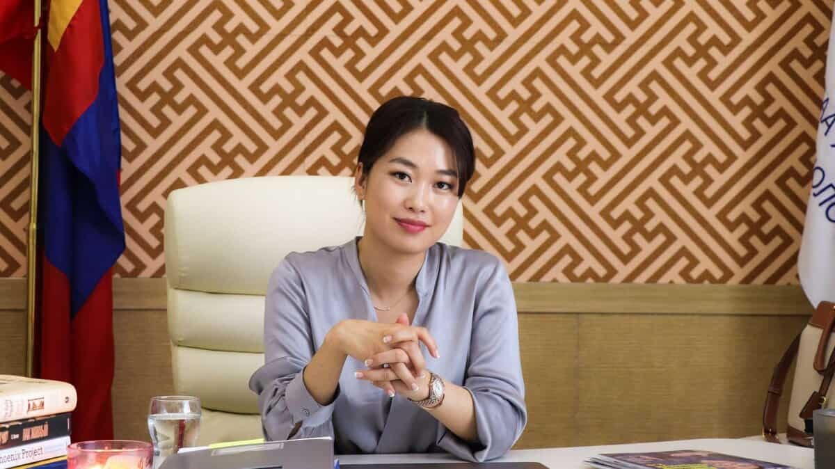 Тайван царайтай эмэгтэй ширээний ард суугаад гараа атгав. Түүний ард геометрийн хээтэй ханын цаас, улсын далбаа харагдана. Ширээн дээр янз бүрийн ном, аяга ус, цаас байна. Тэрээр өөрийн компанийг удирдаж буй энтрепренерийн итгэлийг илэрхийлдэг мөнгөн цамц өмссөн байна. - Business.MN