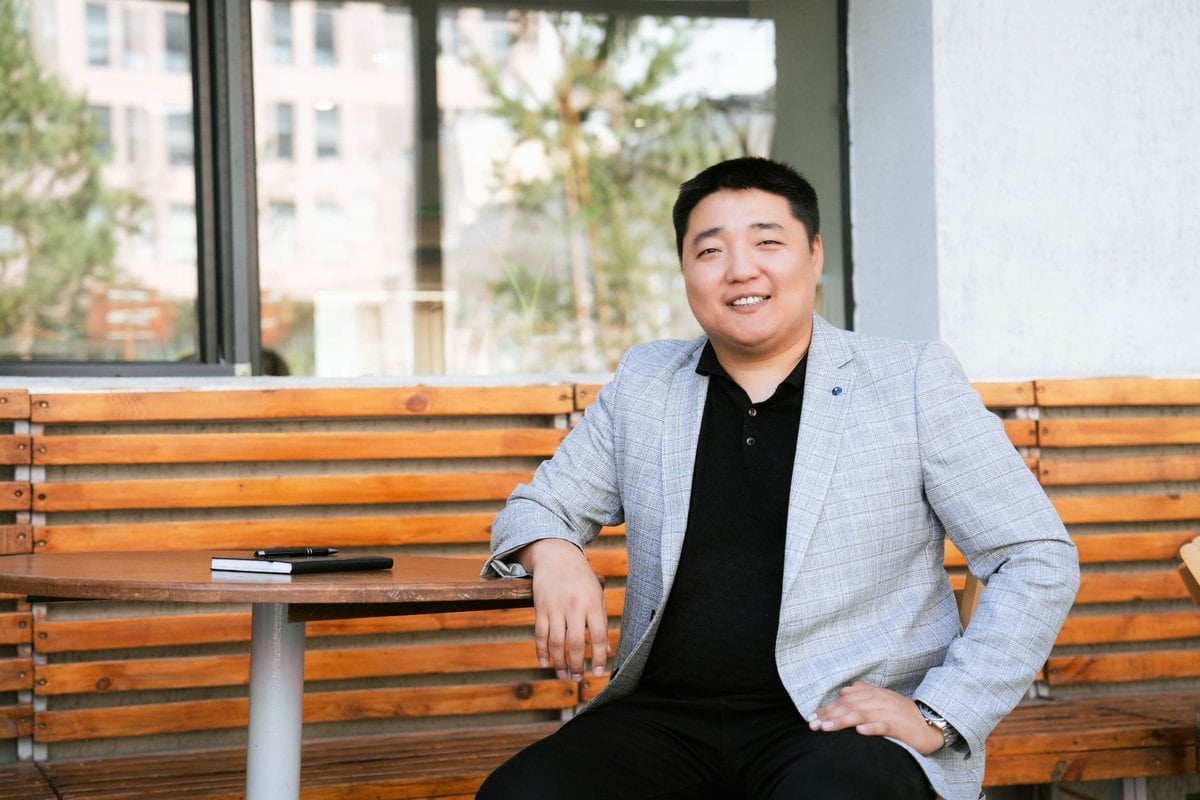 Саарал пиджак, хар цамц өмссөн эрэгтэй гадаа модон вандан сандал дээр суугаад камер руу инээмсэглэв. Түүний хажууд ширээн дээр дэвтэр, утас, магадгүй хөрөнгө оруулалтын асуудлыг хэлэлцэх хэрэгсэл, цаана нь байшин барилга, мод харагдана. - Business.MN