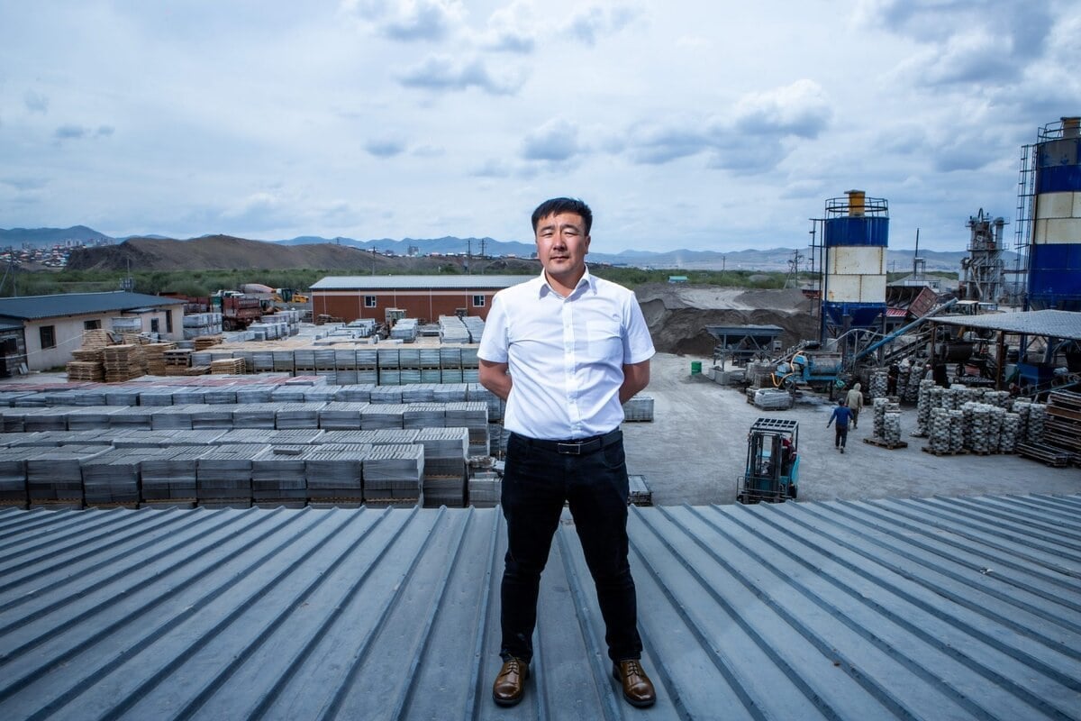 Цагаан цамц, хар өмд өмссөн эр дээвэр дээр үйлдвэрийн талбай, овоолсон материал, үүлэрхэг тэнгэрийн доор уулстай зогсож байна. - Business.MN