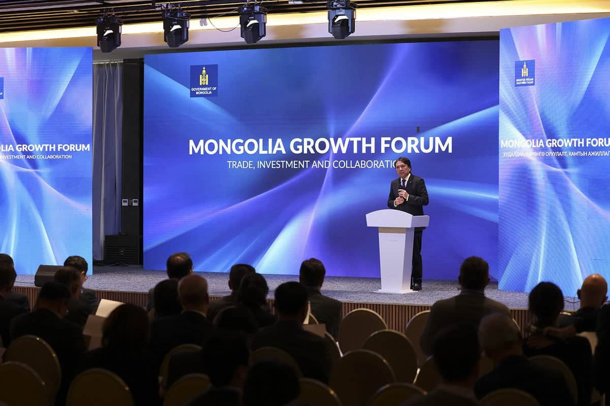 A speaker at a podium addresses an audience at the Mongolia Growth Forum. The backdrop displays the forum's theme: "Trade, Investment and Collaboration," with digital blue graphics and official logos.