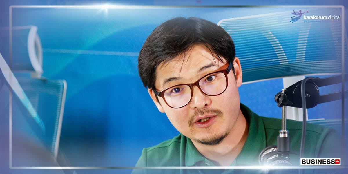 Нүдний шилтэй хүн цэнхэр, мэргэжлийн фонтой микрофон руу ярьж байна. Текст: "Каракорум Дижитал", "Бизнес". - Business.MN