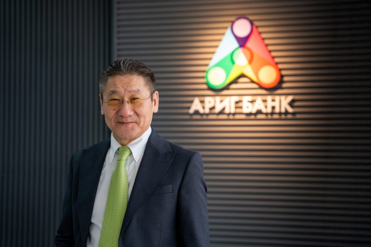 A man in a suit and green tie stands smiling in front of a wall with the colorful Arig Bank logo and Mongolian text.