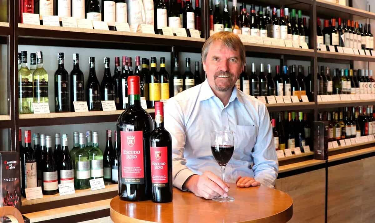 Сахалтай эр дарсны дэлгүүрийн жижиг ширээний ард улаан дарс барин сууж байна. Ширээн дээр хоёр том шил улаан дарс байна. Цаана нь янз бүрийн лонх дарсаар дүүрсэн тавиурууд байна. - Business.MN