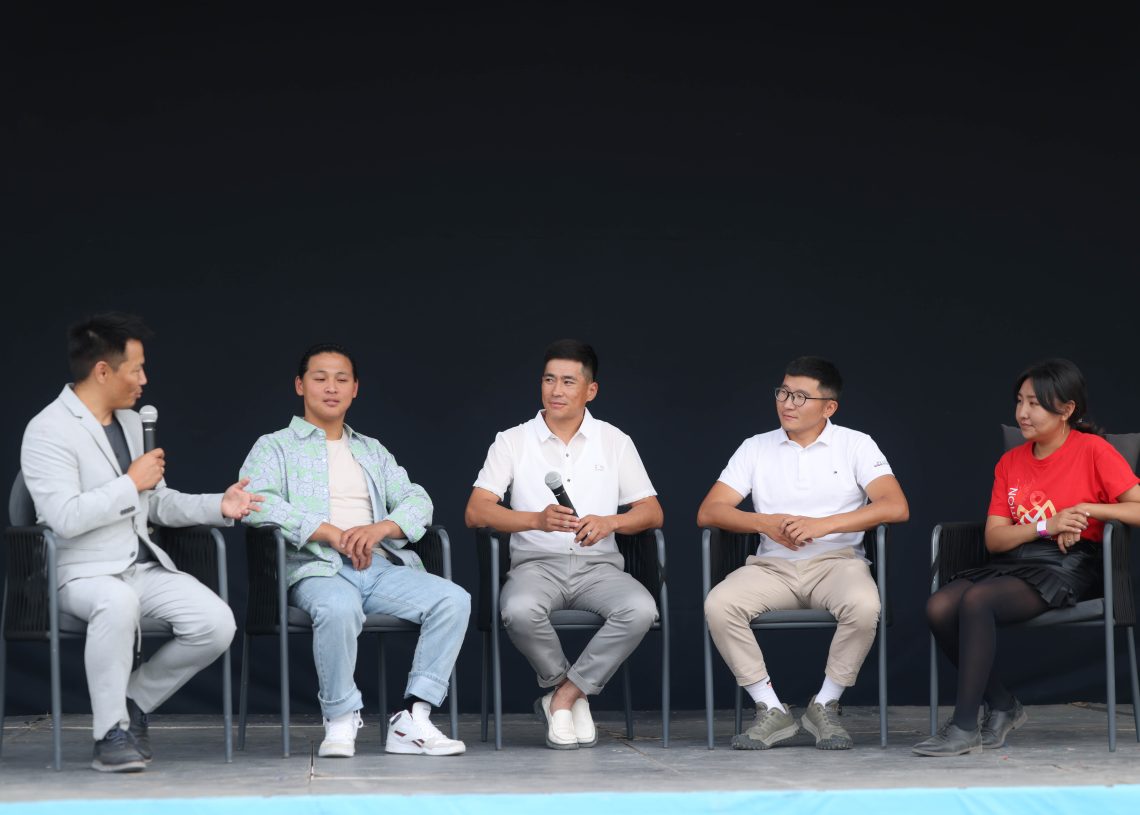 Five people sit in chairs on a stage with microphones. The person on the far left in a gray suit is speaking. The others are casually dressed, including a red shirt, light jackets, and polo shirts. The background is plain and dark.