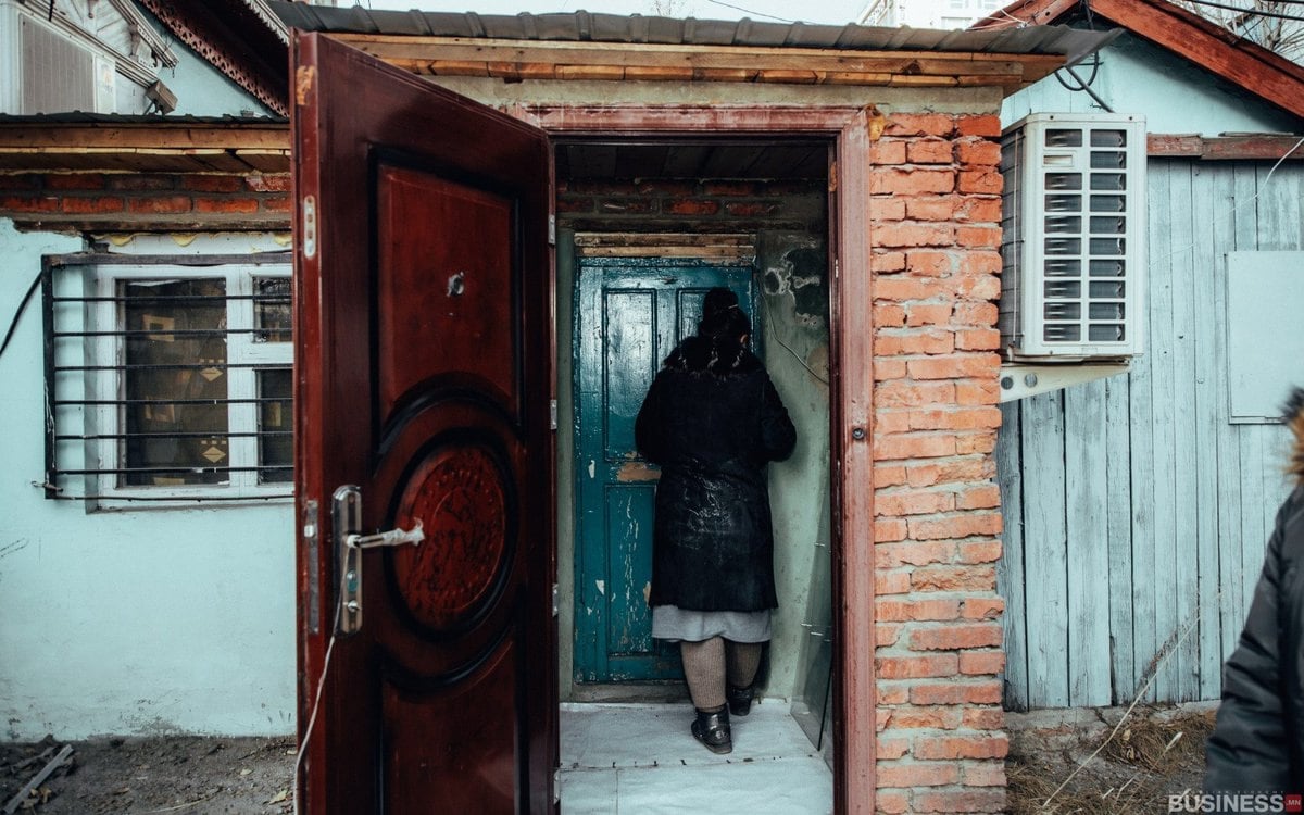 Тоосгон ханатай, оюу хаалгатай, өгөршсөн жижиг барилгын үүдэнд зогсож буй хүн. Хаалга онгорхой, хүн дотогшоо харсан байна. Агааржуулагчийг тоосгон хананд суурилуулсан. - Business.MN