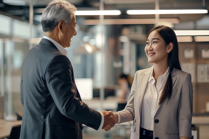 Two business professionals in suits, one older man and one younger woman, smiling and shaking hands in a modern office setting with blurred background and bright lighting.
