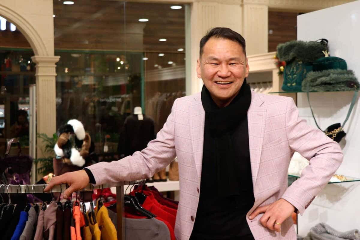 Цайвар ягаан пиджак, хар ороолт өмссөн, амжилттай энтрепренер мэт харагдах эр эргэн тойронд нь янз бүрийн хувцас өмссөн хувцасны дэлгүүрт инээмсэглэж байна. - Business.MN