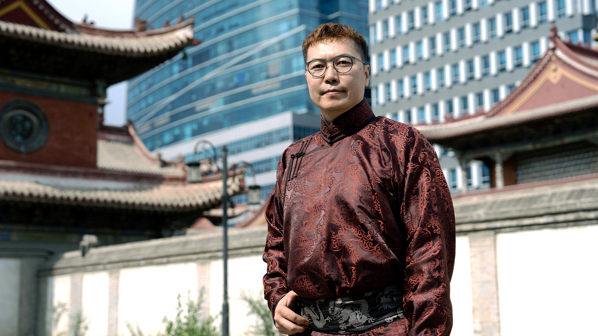 A man wearing traditional patterned maroon Mongolian attire stands outdoors, with historical and modern buildings visible in the background.