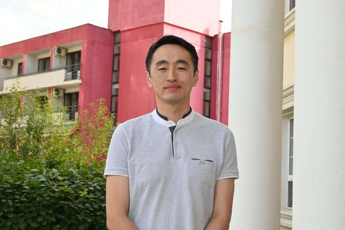 Богино хар үстэй, цайвар саарал поло цамц өмссөн эрэгтэй цагаан баганын өмнө зогсож, арын дэвсгэр дээр улаан фасадтай барилга байна. Зургийн зүүн талд ногоон навч харагдана. Тэрээр бизнес эрхэлдэг хүний тайван байдлыг харуулсан төвийг сахисан харцаар камер руу харж байна. - Business.MN