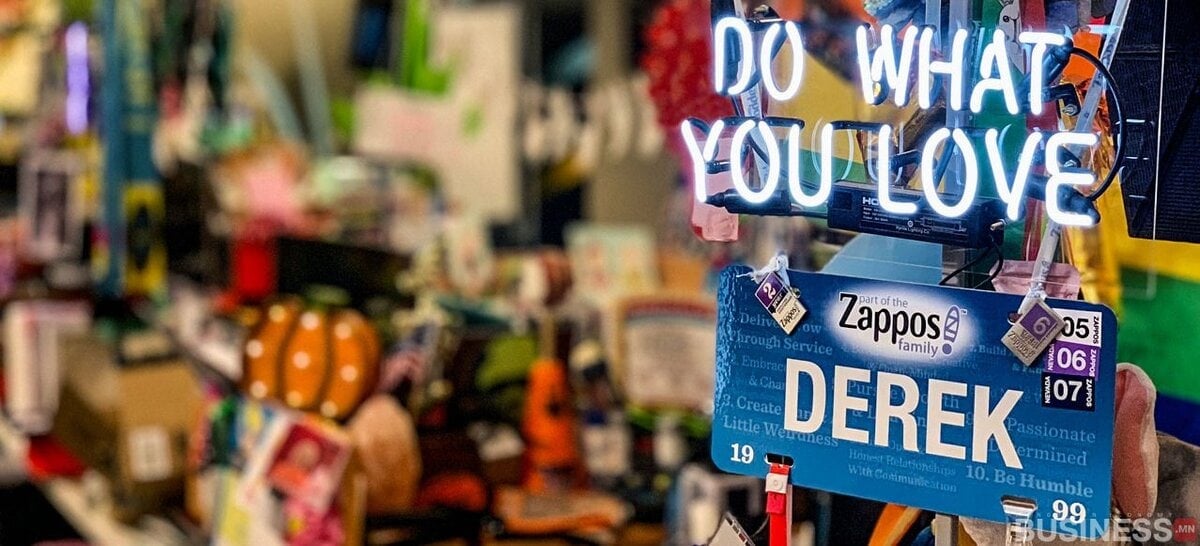 "ХАЙРТАЙ ЗҮЙЛЭЭ ХИЙ" гэсэн неон самбар, янз бүрийн зүйлээр хүрээлэгдсэн "DEREK" гэсэн шошго бүхий эмх замбараагүй ажлын талбар нь энтрепренерийн бужигнааныг илэрхийлдэг. - Business.MN