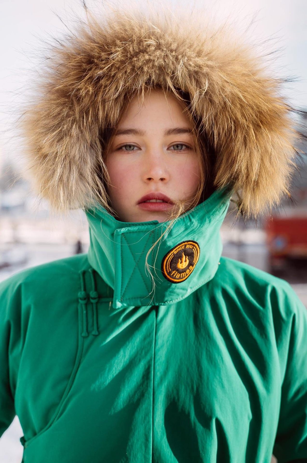 A young person in a green winter coat with a large, furry hood stands outside in a snowy setting. Their face is framed by the hood, and a yellow patch is visible on the coat. The background is blurred.
