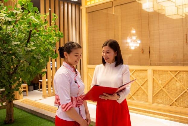 Хоёр эмэгтэй дотор нь онгорхой улаан хавтсыг хараад зогсож байна. Зүүн талд байгаа үсээ сүлжсэн, улаан өнгийн цагаан өнгийн цамц өмссөн эмэгтэй инээмсэглэнэ. Баруун талд байгаа эмэгтэй, мөр хүртэл урт үстэй, цагаан, улаан хувцастай энтрепренер байж магадгүй хавтас руу дохиж байна. - Business.MN