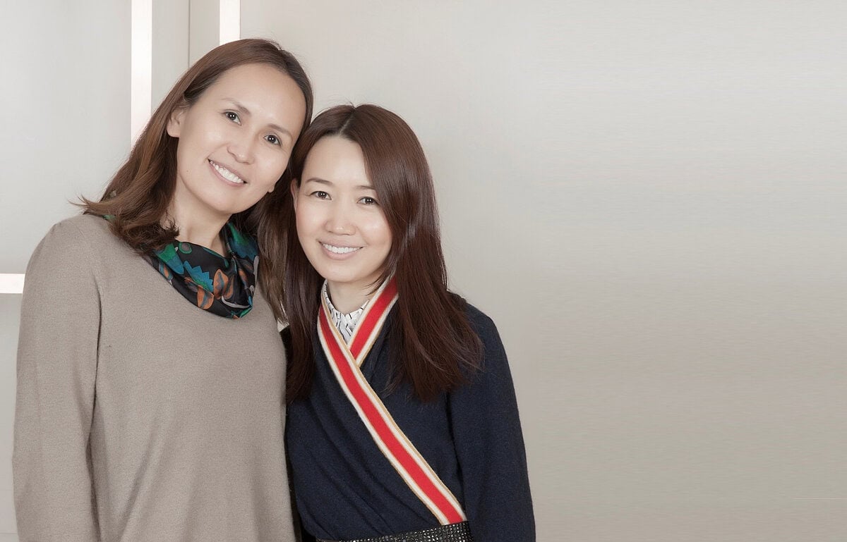 Хоёр эмэгтэй ойрхон зогсож, цайвар өнгийн дэвсгэр дээр камер руу инээмсэглэж байна. Нэг нь шаргал ноосон цамц өмссөн, нөгөө нь судалтай ороолттой бараан өнгийн цамцтай. - Business.MN