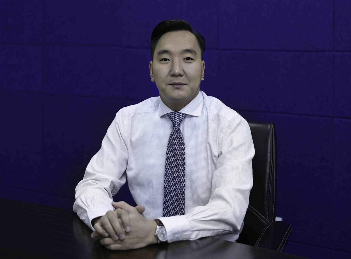 Цагаан даашинзтай цамц, хээтэй зангиа зүүсэн эр бараан модон ширээний ард гараа атгасан сууж байгаа нь энтрепренерийн нухацтай зан чанарыг илтгэнэ. Тэрээр богино хар үстэй бөгөөд гүн цэнхэр дэвсгэр дээр шууд камер руу харж байна. - Business.MN