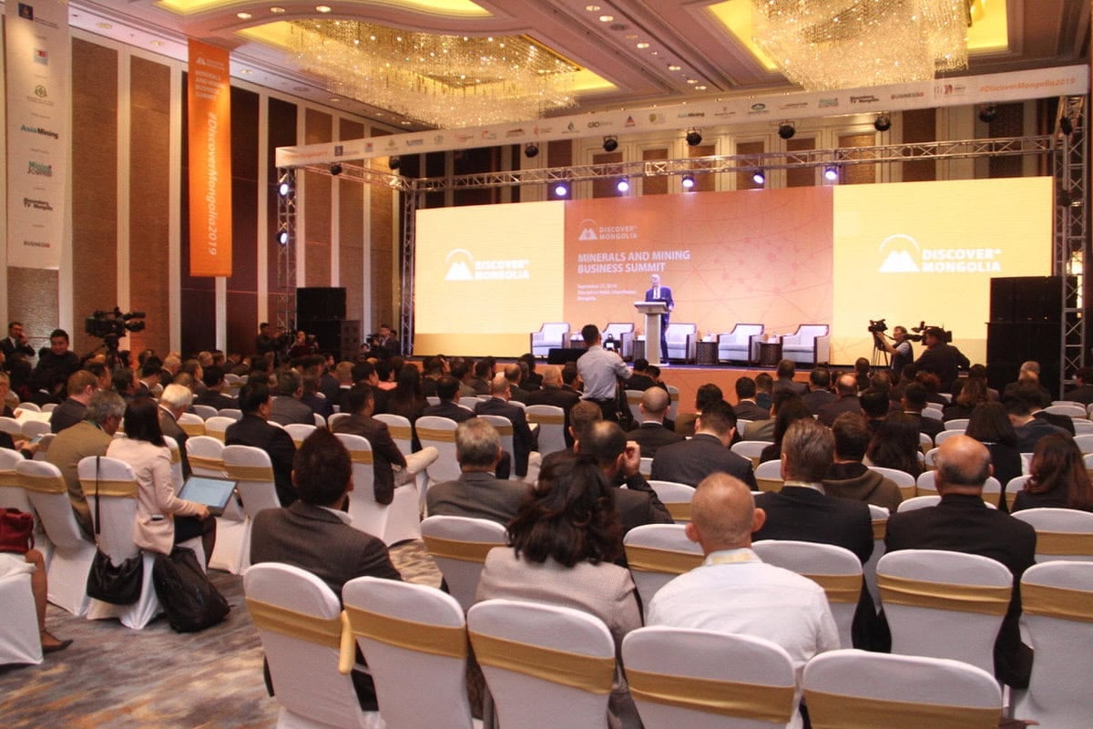 Эзлэгдсэн сандал бүхий том хурлын танхим. "Дисковер Монголиа: Эрдэс баялаг, уул уурхайн бизнесийн дээд хэмжээний уулзалт" гэсэн бичигтэй тод дэлгэцийн өмнө илтгэгч тайзан дээр зогсож байна. Оролцогчид илтгэлд анхаарлаа хандуулдаг. - Business.MN