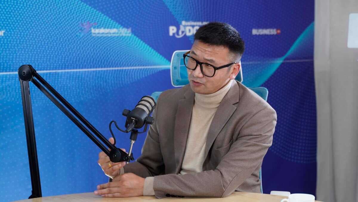 Нүдний шил, цайвар өнгийн яст мэлхийтэй бор пиджак өмссөн хүн индэр дээр суурилуулсан микрофон руу ярьж байна. Арын дэвсгэр нь бизнесийн подкаст эсвэл ярилцлагын тохиргоог санал болгож буй текст бүхий цэнхэр геометрийн загвартай. Ширээн дээр кофены аяга байна. - Business.MN