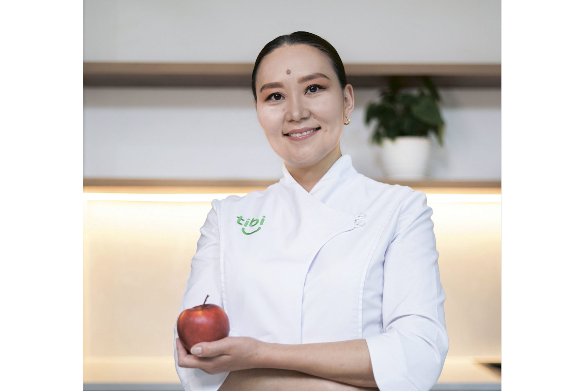 Орчин үеийн гал тогооны өрөөнд ногоон компани логотой хатгамал бүхий цагаан тогоочийн цув өмссөн эмэгтэй баруун гартаа улаан алим барин инээмсэглэн зогсож байна. Цаана чинь ваартай ургамал байна. - Business.MN