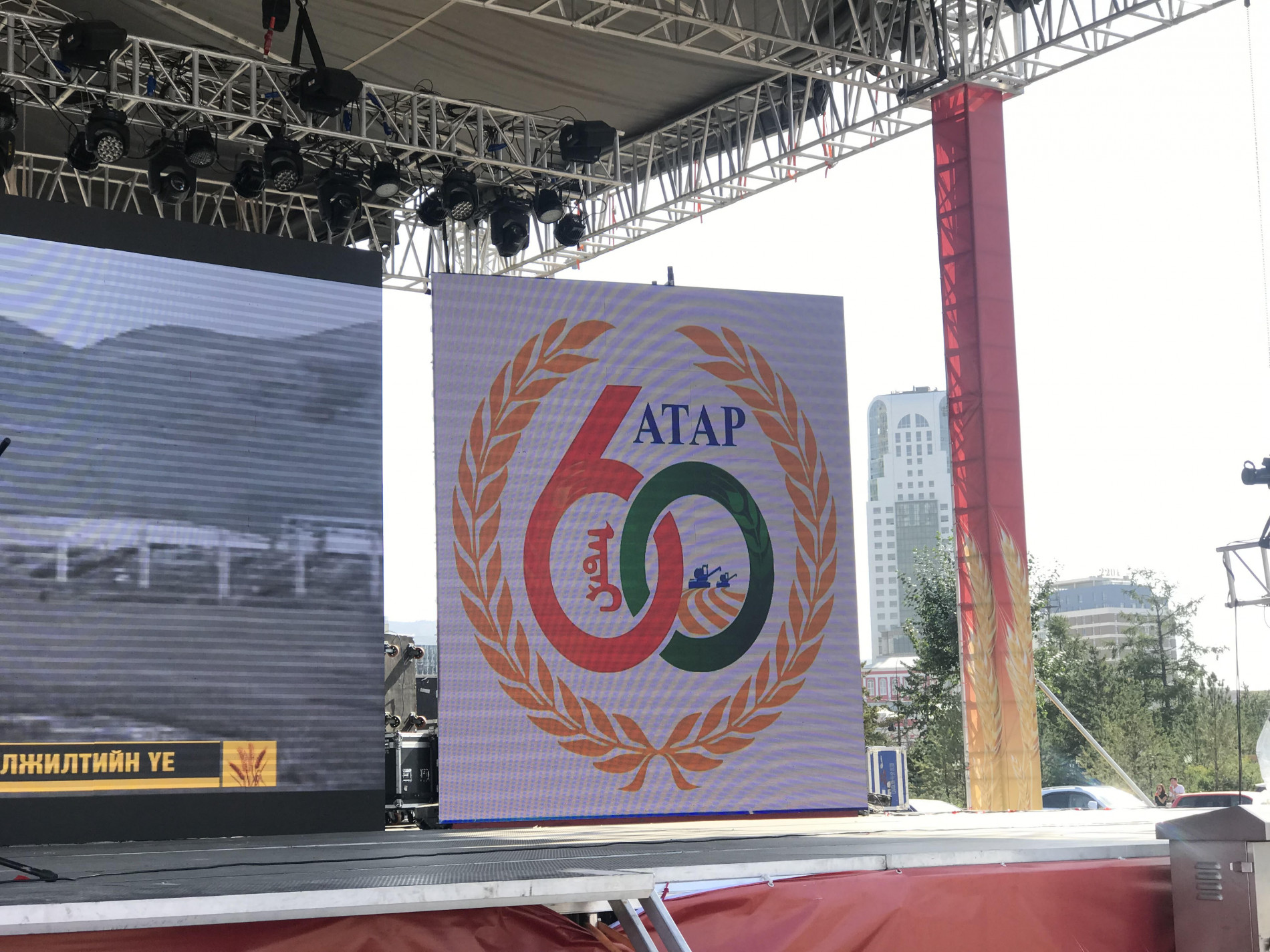 Лаврын алтан хэлхээгээр хүрээлэгдсэн 60 жилийн ойн бэлгэ тэмдгийг харуулсан том дэлгэц бүхий гадаа тайз. Арын дэвсгэр нь хотын дүр төрхийг агуулдаг. - Business.MN