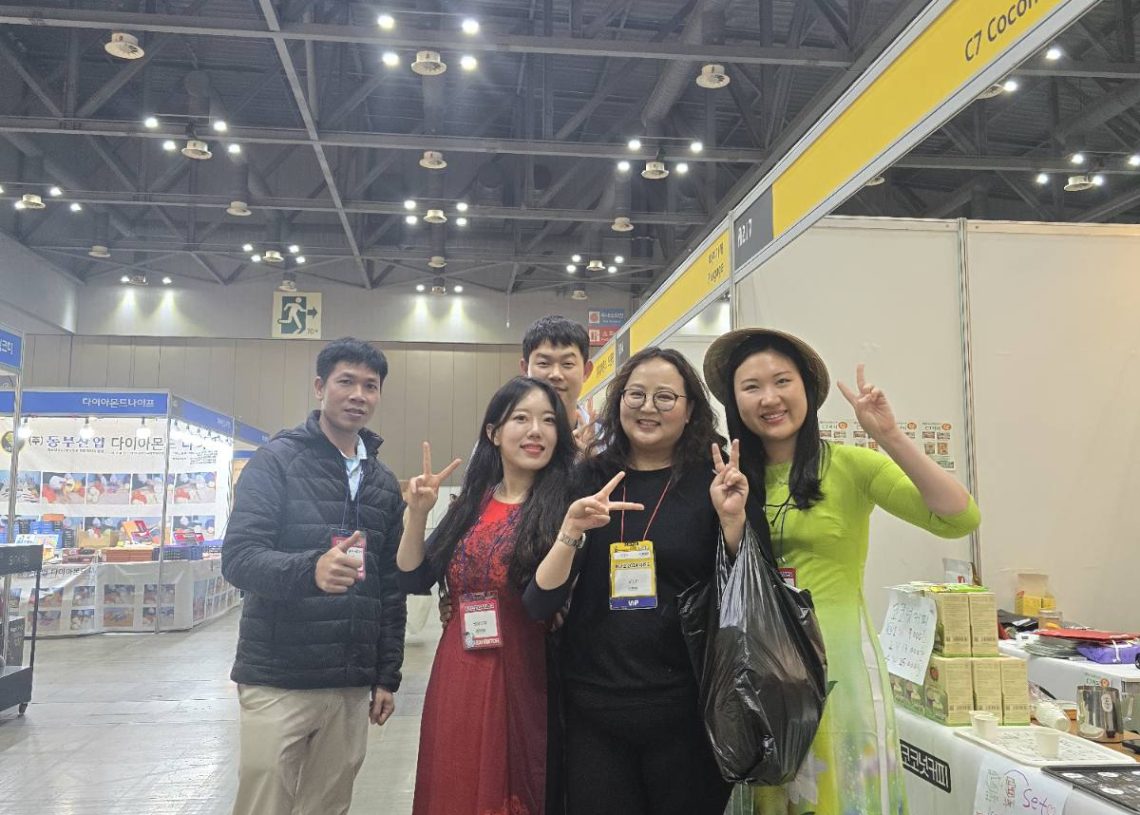 Five people are standing and smiling in an indoor exhibition hall. They are next to a display booth with various items. Two women are wearing traditional dresses. The group is making peace signs with their hands.