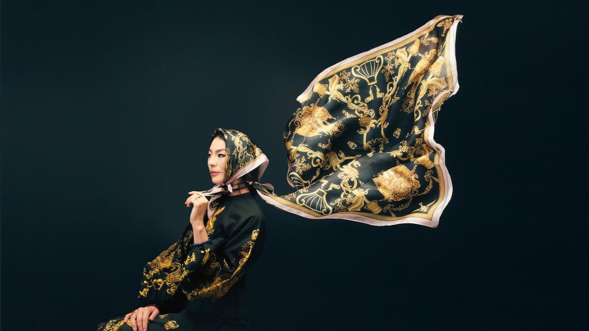 Хар даашинз, гоёмсог хар алтан торгон ороолт өмссөн эмэгтэй бараан дэвсгэр дээр сууж байна. Ороолт нь салхинд баригдсан мэт түүний араас огцом урсдаг. - Business.MN