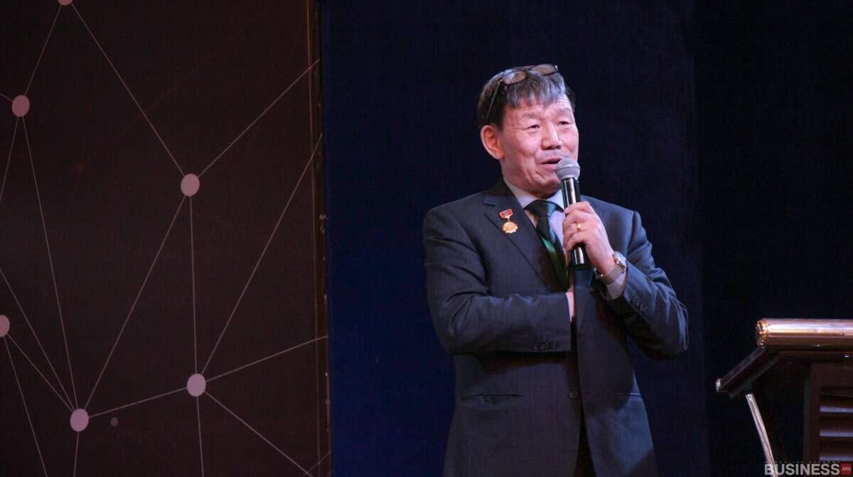 Костюм өмссөн эр тайзан дээр микрофон руу ярьж, ард нь хээтэй дэвсгэртэй, компанийнхаа ирээдүйн талаар ярилцаж байна. - Business.MN