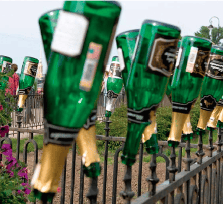 Гадна цэцэрлэгт хүрээлэнгийн хар төмөр хашаан дээр алтан тугалган цаас бүхий хөмөрсөн ногоон шилэн лонхны эгнээ байрлуулсан бөгөөд урд талд нь цэцэг, ард нь мод харагдаж байна. - Business.MN