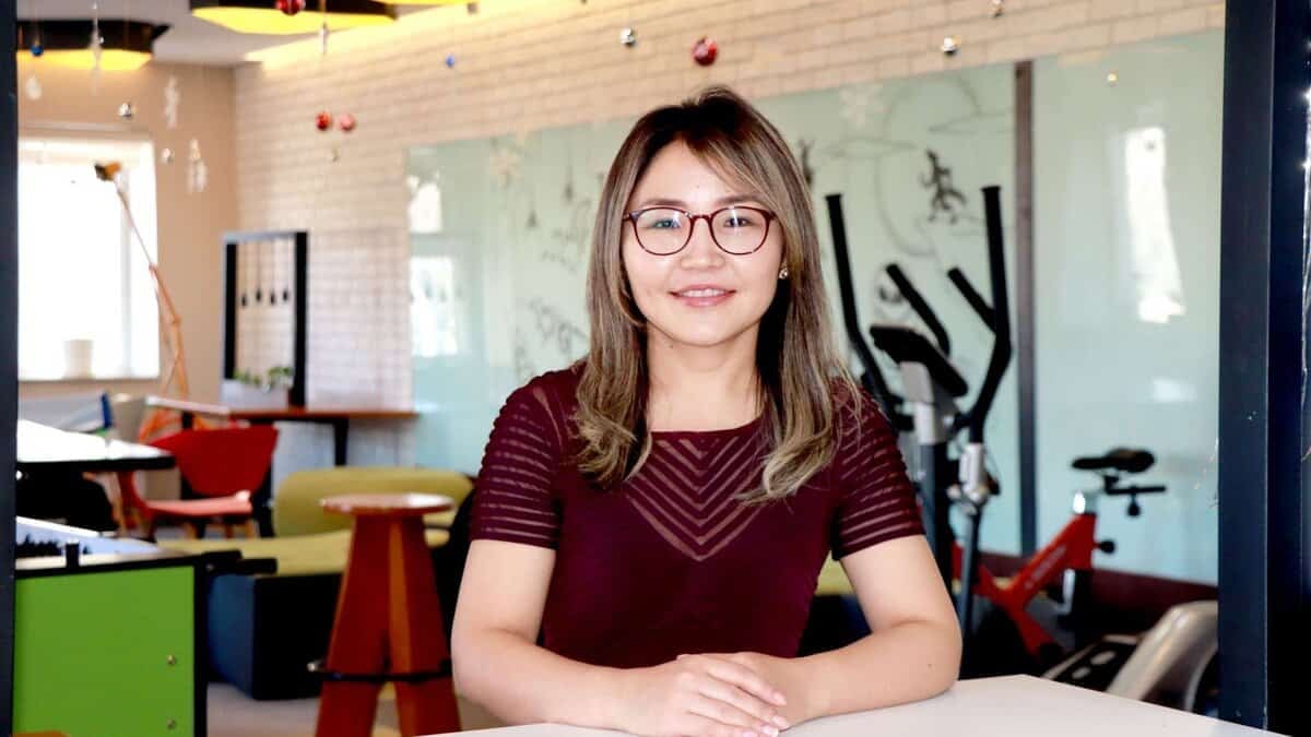 Нүдний шилтэй, урт үстэй эмэгтэй орчин үеийн оффисын өрөөнд хүрэн улаан даашинз өмссөн итгэлтэй зогсож байна. Арын дэвсгэр дээр өнгөлөг тавилга, дасгалын тоног төхөөрөмж, гоёл чимэглэлийн элементүүд байдаг. - Business.MN