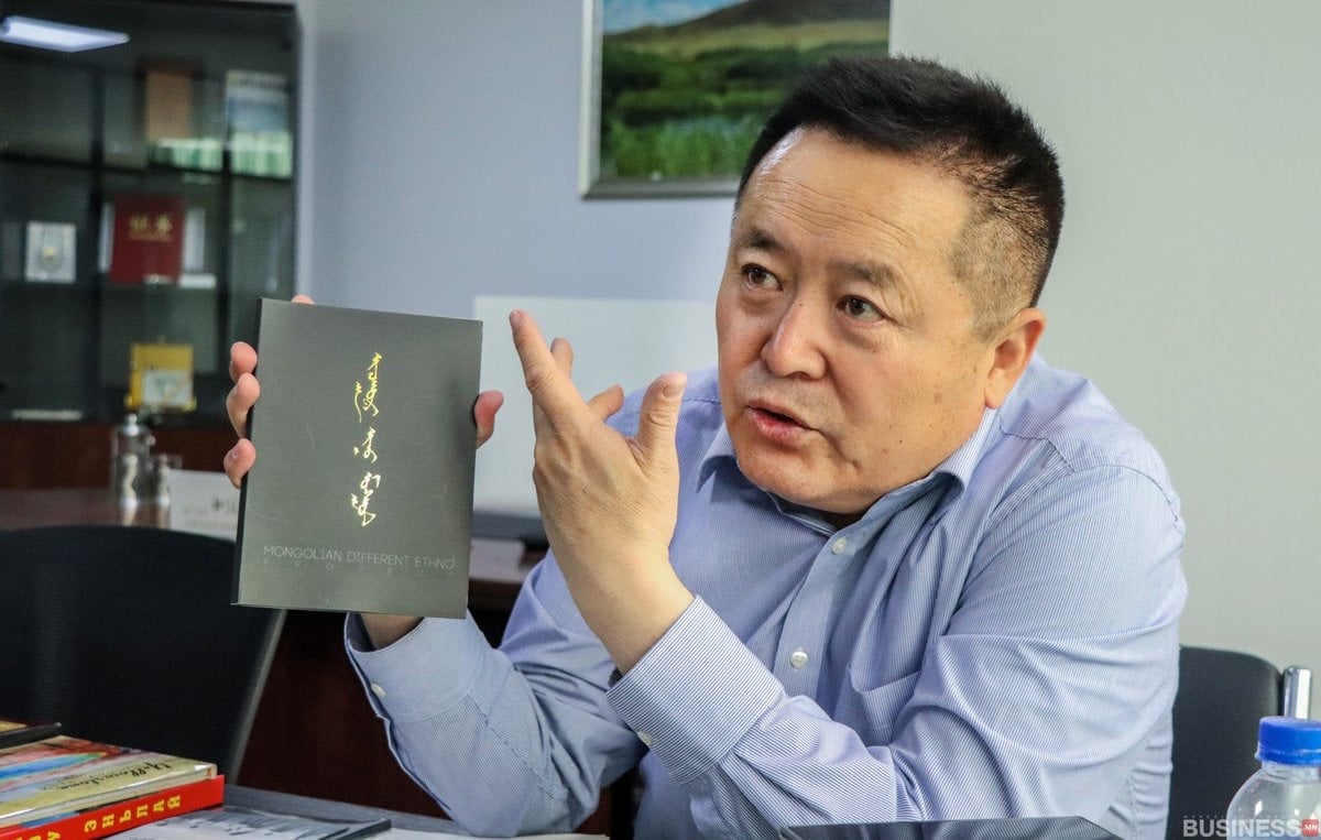Цэнхэр цамцтай эр бараан хавтастай "Монгол угсаатан" нэртэй номыг барин дохиж байна. Тэр ширээний ард сууж, урд нь өөр хэдэн ном байна. Ар талд нь шүүгээ болон жаазтай зураг байна. - Business.MN
