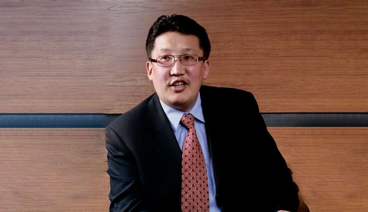 Богино хар үстэй, нүдний шил, сахалтай, хар костюм, цайвар цэнхэр цамц, хээтэй улаан зангиа зүүсэн эр модон хавтангийн дэвсгэр дээр сууж байна. Тэрээр компанийнхаа талаар ярьж эсвэл ярьж байгаа бололтой. - Business.MN