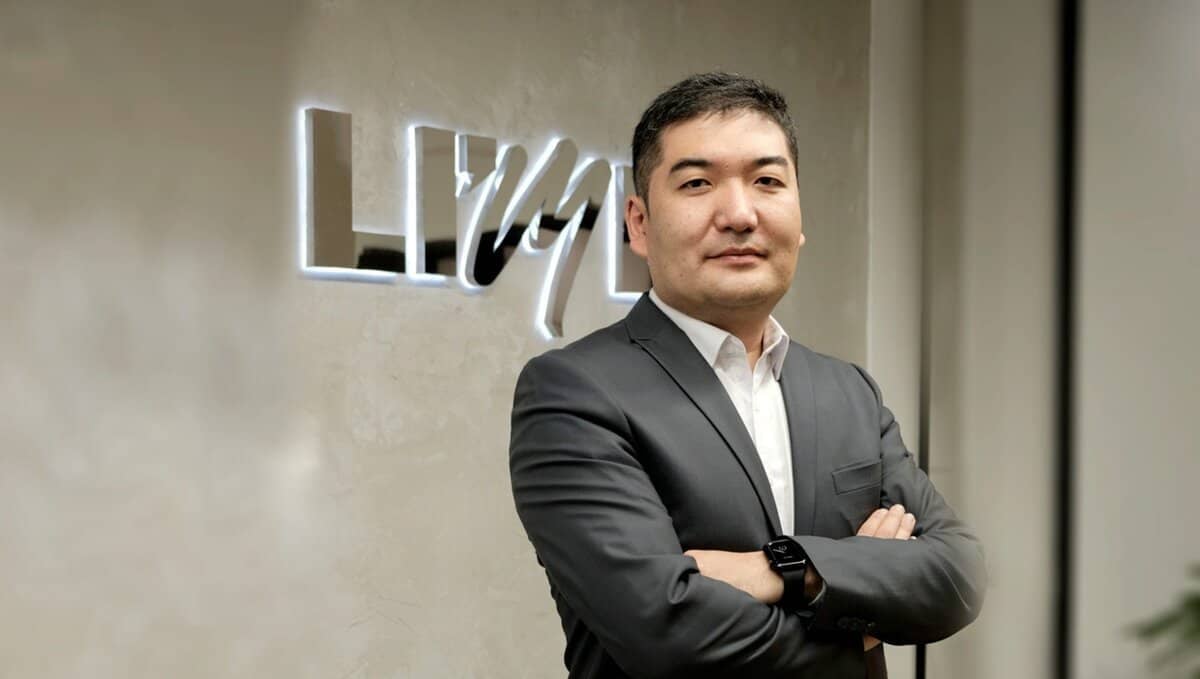 Цагаан цамцтай саарал костюмтай хүн "LIV" гэсэн бичигтэй хананы өмнө гараа зөрүүлэн зогсож байна. Энтрепренер хүн богино хар үстэй, зүүн бугуйндаа ухаалаг цаг зүүсэн байна. Арын дэвсгэр нь орчин үеийн, төвийг сахисан өнгө аястай интерьерийг харуулдаг. - Business.MN