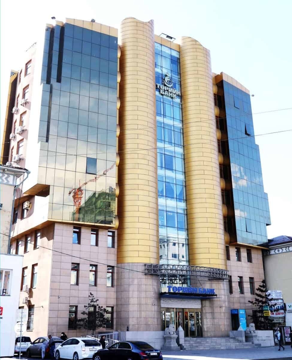 Гадна талын гэрэл цацруулагч шилтэй, алтлаг өнгийн босоо өргөлт бүхий олон давхар барилга тод харагдаж байна. Орцны дээд талд англи бус бичгээр бичигдсэн бөгөөд энэ нь бизнесийн компанийн төв байр байж магадгүй гэсэн санаа юм. Урд талд нь машинууд зогсож, гудамжинд явган зорчигч, тээврийн хэрэгсэл идэвхтэй байдаг. - Business.MN