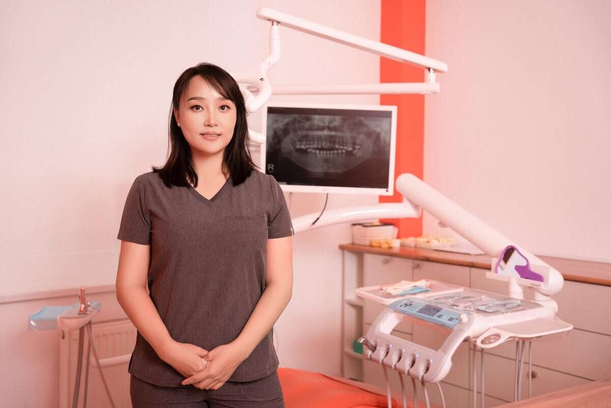 A dentist in scrubs stands in a dental office with a dental chair, equipment, and an X-ray displayed on a monitor. The room is well-lit with orange accents.