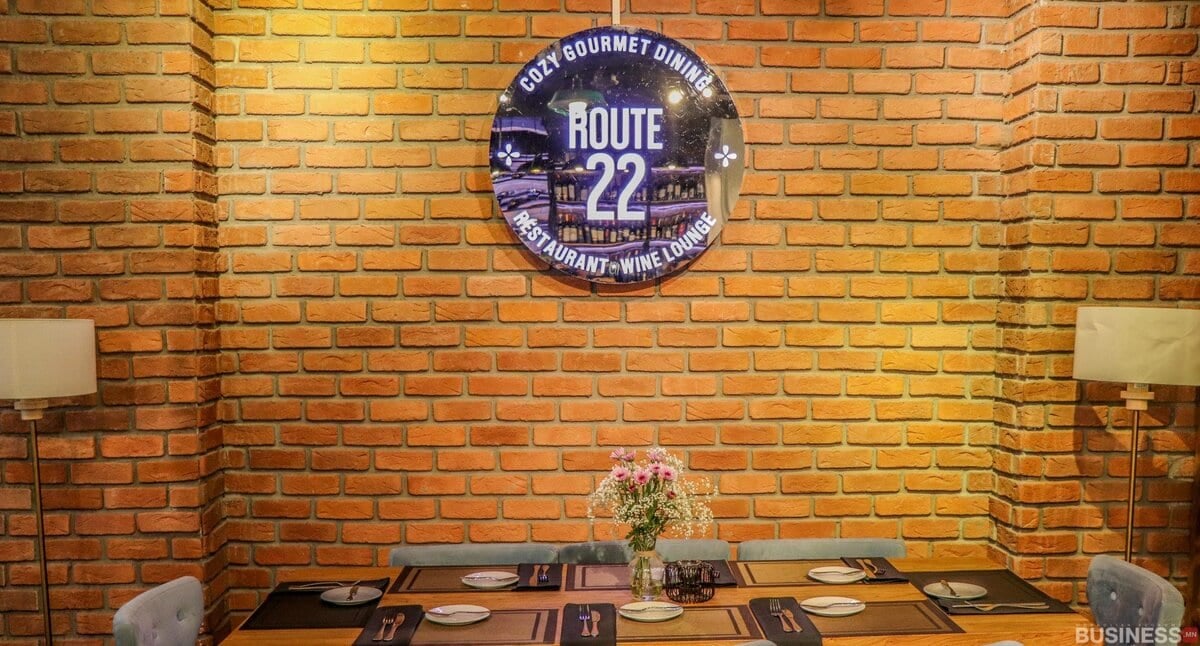 Тоосгон хананд наасан зургаан хүний модон ширээ бүхий тав тухтай хоолны газар. Ханан дээр "Маршрут 22" гэсэн дугуй хэлбэртэй тэмдэг бүхий тансаг зоогийн газар, дарсны танхимыг сурталчилсан байна. Шалны хоёр чийдэн нь ширээний хажууд байгаа нь халуун дулаан уур амьсгалыг бүрдүүлдэг. - Business.MN