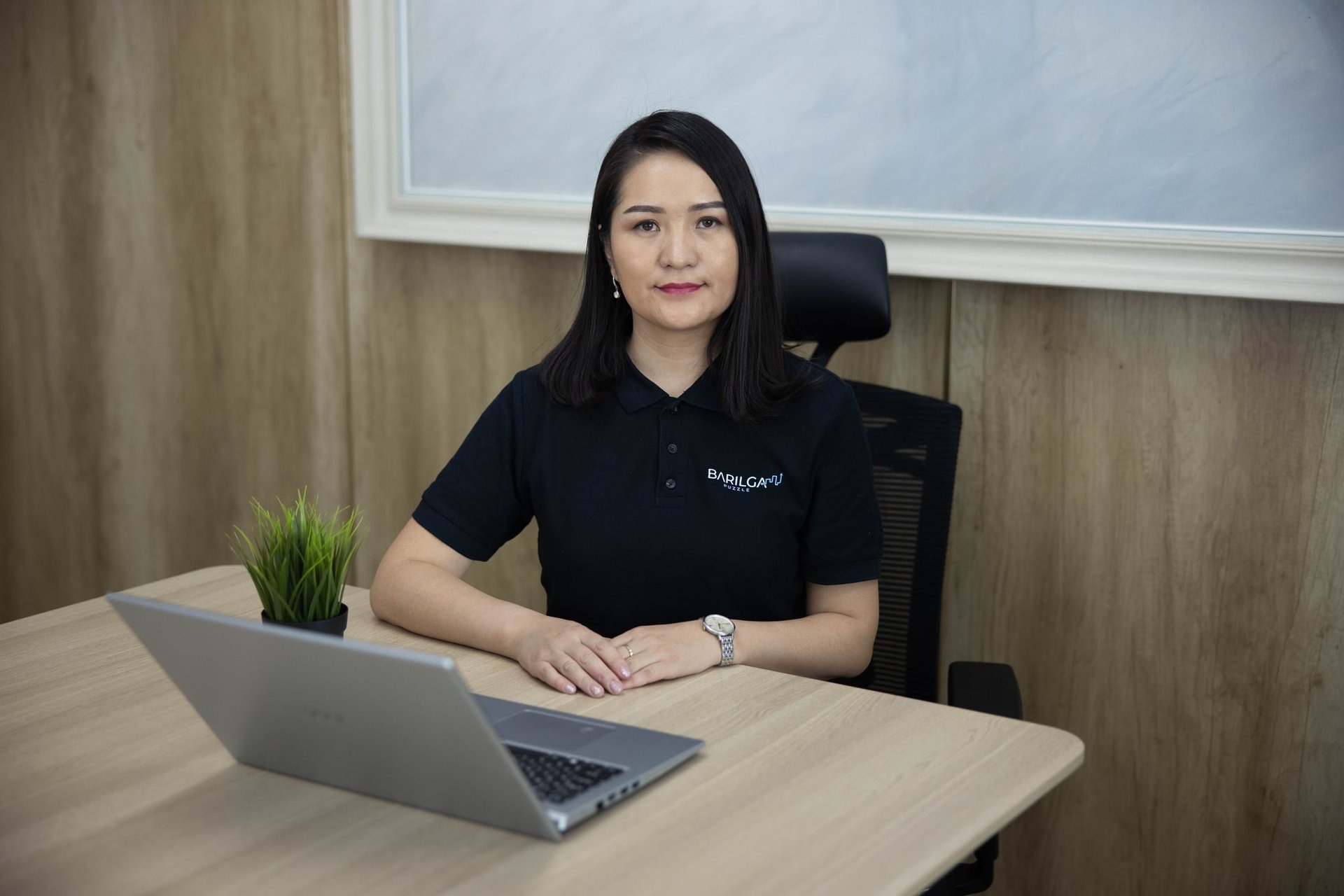 Шулуун, мөр хүртэл урт хар үстэй эмэгтэй мөнгөн зөөврийн компьютер, жижиг савтай ургамал бүхий модон ширээний ард сууж байна. Компанийхаа лого бүхий хар поло цамц өмссөн энтрепренер нь модон хавтангаар хүрээлэгдсэн бөгөөд цаана нь том жаазтай зураг байдаг. - Business.MN