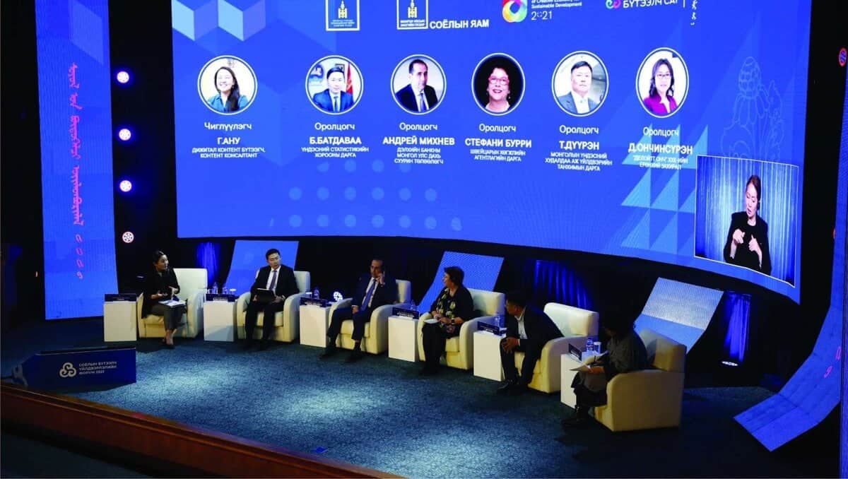 Зургаан илтгэгч, дөрвөн эрэгтэй, хоёр эмэгтэй хүний зураг, нэрийг харуулсан том дижитал дэлгэц бүхий тайзан дээр сууж байна. Үзэгчдийн талбай бүдэгхэн гэрэлтэй байхад тайз гэрэлтүүлэг сайтай. Баруун талд дохионы хэлний орчуулагч зогсож байна. - Business.MN