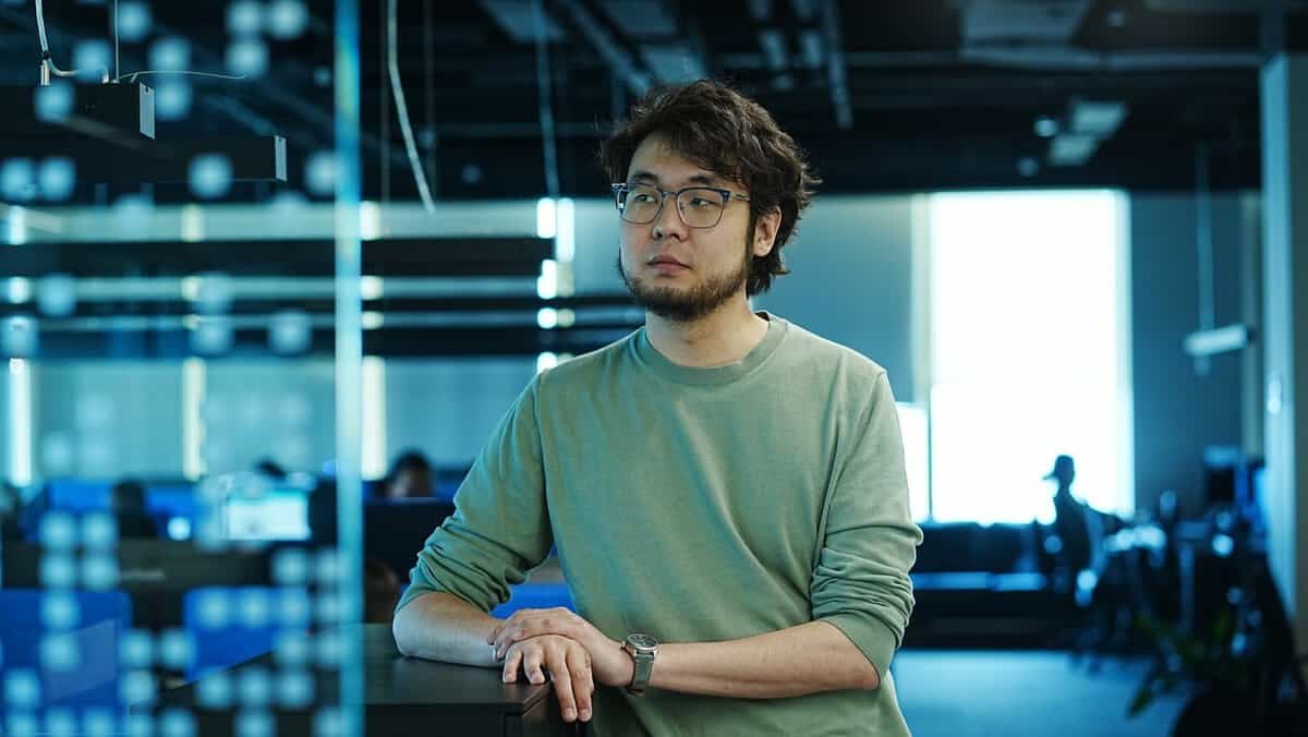 Нүдний шилтэй, сахалтай, ногоон ноосон цамц өмссөн хүн орчин үеийн, цэнхэр өнгийн чимэглэлтэй, задгай шалтай дизайнтай оффисын орчинд тайван зогсож байна. Нэг гараа ширээн дээр тавин, энтрепренер алсыг харж, дараагийн бизнесээ эргэцүүлэн бодож байна. - Business.MN