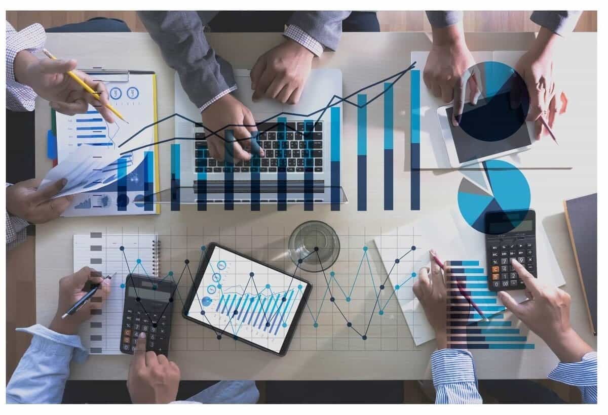 Зөөврийн компьютер, таблет, тооны машин, графикаар хүрээлэгдсэн ширээний ард бизнес төсөл дээр хамтран ажиллаж буй бизнес эрхлэгчдийн дээрээс доошоо харагдах байдал. График болон өгөгдлийн дүрслэл нь дижитал байдлаар давхардсан бөгөөд энэ нь компанид дүн шинжилгээ хийх, багаар ажиллах чадварыг илэрхийлдэг. - Business.MN