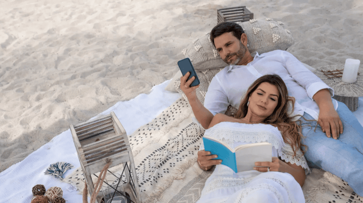 A couple relaxes on a sandy beach; the woman is lying on the man’s chest, reading a book, while the man looks at his phone. They are on a blanket with cushions and decorative wooden lanterns nearby.