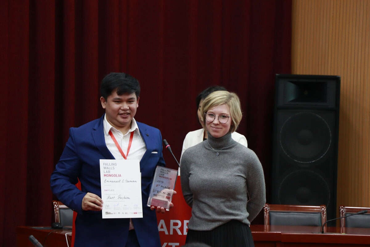 Хоёр хүн тайзан дээр зогсож байна. Зүүн талд байгаа хүн шагнал, гэрчилгээтэй. Тэд хоёулаа инээмсэглэж байна. Арын дэвсгэр дээр улаан хөшиг, том чанга яригч харагдаж байна. - Business.MN