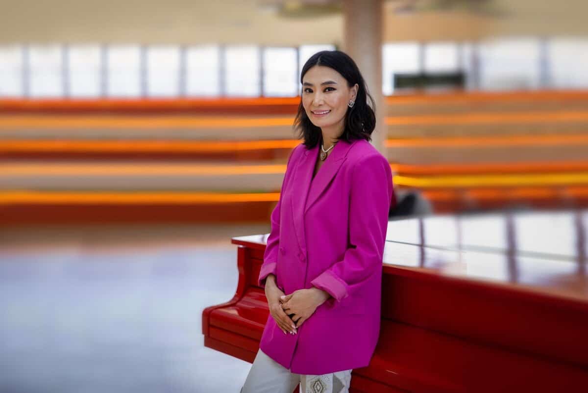 Өөртөө итгэлтэй энтрепренер шиг тод ягаан пиджак өмссөн эмэгтэй улаан төгөлдөр хуурын дэргэд зогсож байна. Тэр мөр хүртэл урт хар үстэй, инээмсэглэж байна. Арын дэвсгэр дээр бүдгэрсэн эгнээ бүхий модон суудлууд байгаа нь орчин үеийн, задгай орон зай, магадгүй концертын танхим эсвэл арга хэмжээ болох газрыг санал болгож байна. - Business.MN