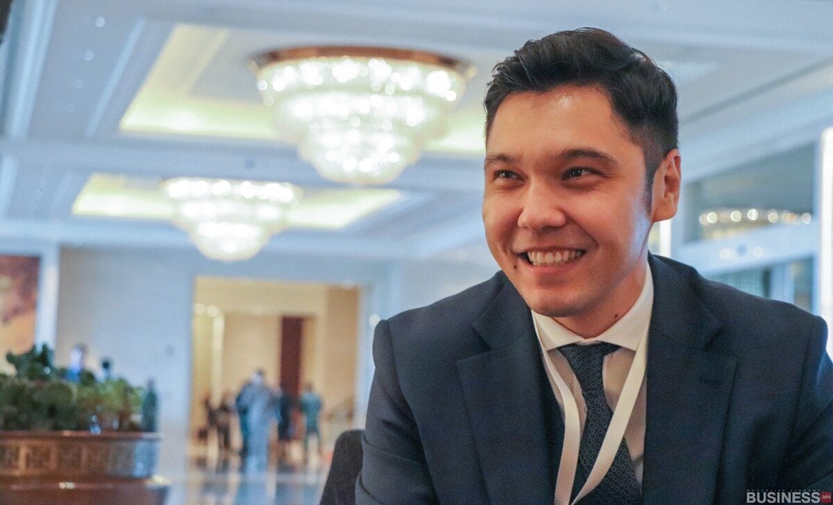 Костюм өмссөн эрэгтэй лааны суурьтай гэрэлтэй өрөөнд сууж байхдаа инээмсэглэж байна. Тэр зангиа зүүж, үнэмлэхний оосортой. Арын дэвсгэр нь зөөлөн бүдгэрч, алсад байгаа хүмүүстэй өргөн талбайг харуулж байна. - Business.MN