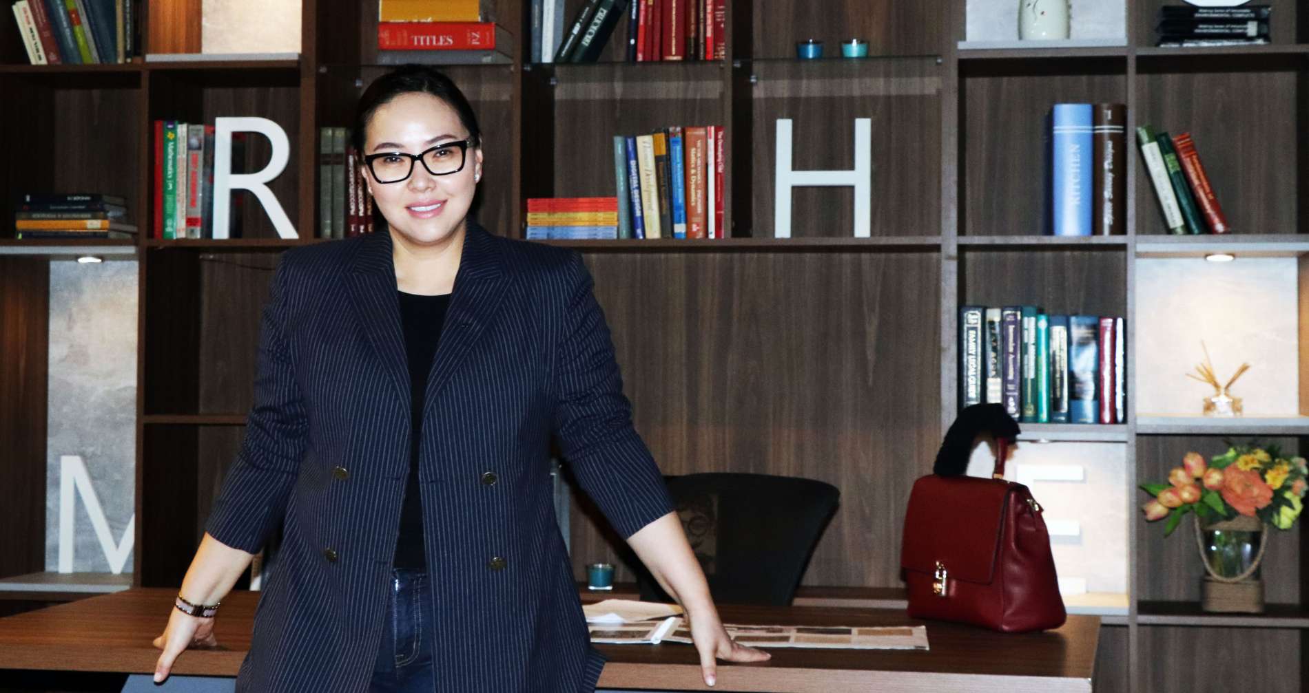 Нүдний шил, бараан пиджак өмссөн хүн ном, гоёл чимэглэлийн зүйлсээр дүүрсэн модон номын тавиурын өмнө зогсож байна. Тэдний хажууд ширээн дээр улаан цүнх сууж байна. - Business.MN