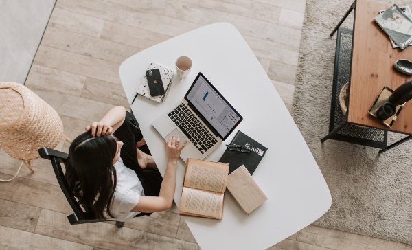 Урт үстэй хүн цагаан ширээний ард суугаад зөөврийн компьютер дээр ажиллаж байна. Ширээн дээр задгай дэвтэр, хаалттай ном, ухаалаг утас, кофены аяга бас байдаг. Энэ үзэгдэл нь модон шал, хивсэнцэртэй, тухтай өрөөнд байдаг. Энтрепренер ажилдаа анхаарлаа хандуулж байгаа бололтой. - Business.MN