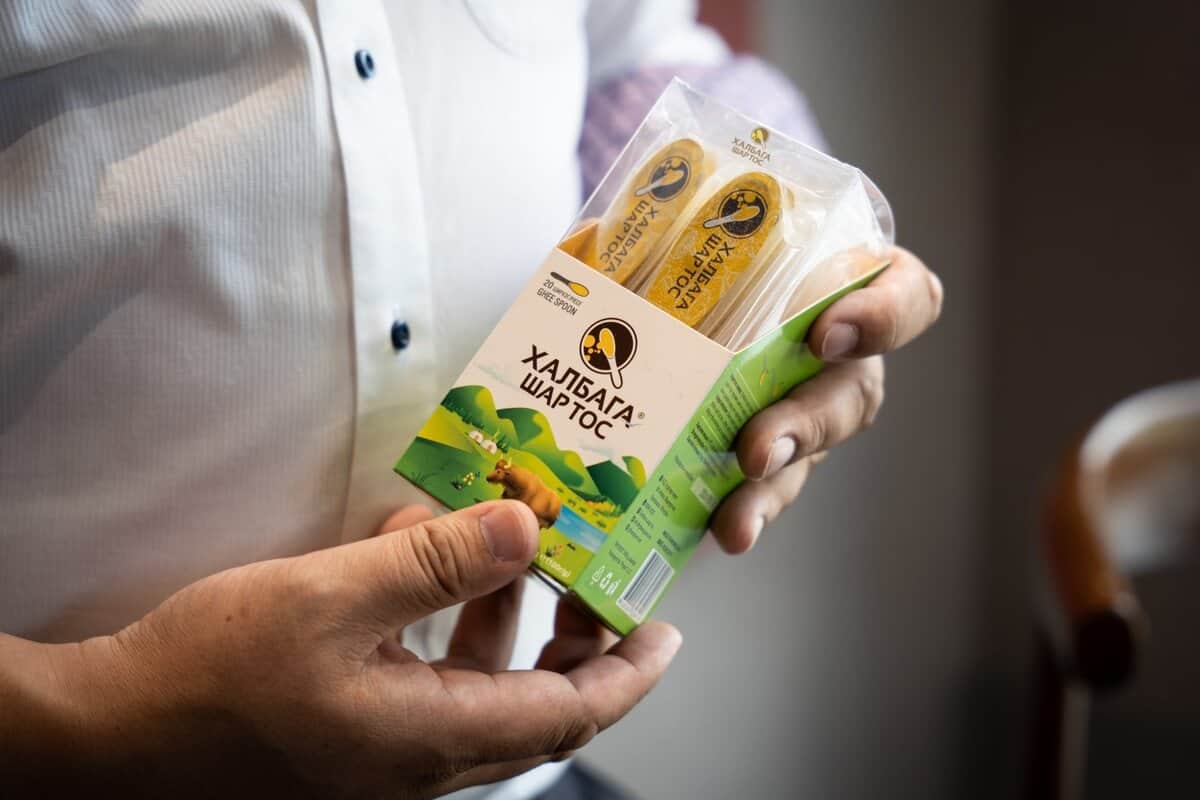 Цагаан цамц өмссөн хүн савласан бүтээгдэхүүн барьж байна. "Дэлхийд таашаагдах Монгол амт" гэсэн үг хэллэг бүхий фермийн өнгө үзэмжээр чимэглэсэн сав баглаа боодол нь хоол хүнс эсвэл хөнгөн зууш агуулж байгааг харуулж байна. Багц дээр англи бус хэл дээрх бичвэр харагдаж байгаа нь түүний чамин гарал үүслийг илтгэнэ. - Business.MN