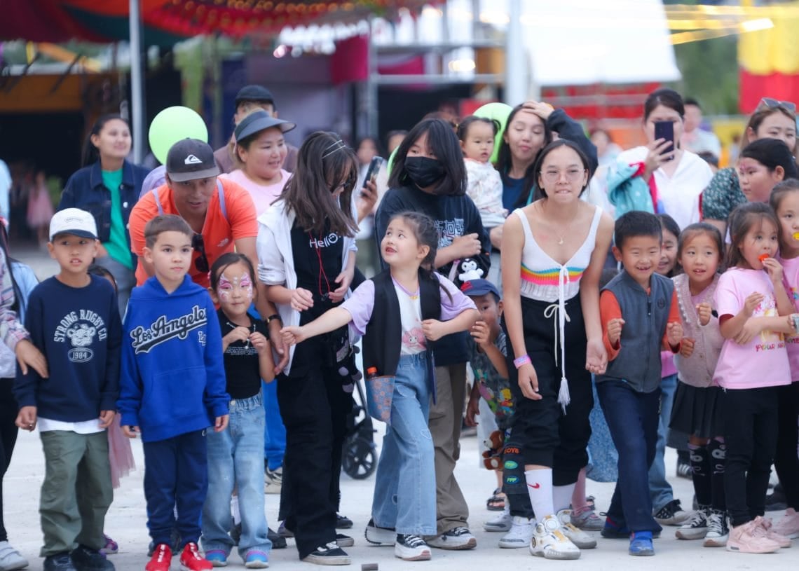 A diverse group of children and adults stands close together, smiling and eagerly watching an unseen event. The background features colorful carnival tents and decorations. Some kids are holding balloons, and the atmosphere is festive and cheerful.