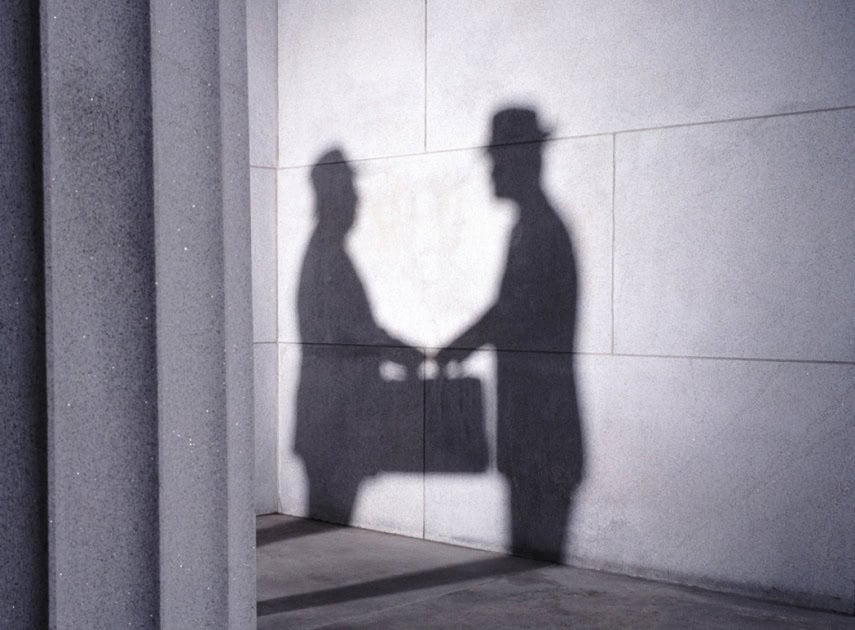 Малгай өмссөн хоёр энтрепренеровын сүүдэр хананд гар барьж, нэг нь цүнх барьдаг. Арын дэвсгэр нь гөлгөр, цайвар өнгийн хана бөгөөд зүүн талын босоо тулгуур баганагаар хэсэгчлэн бүрхэгдсэн байдаг. - Business.MN