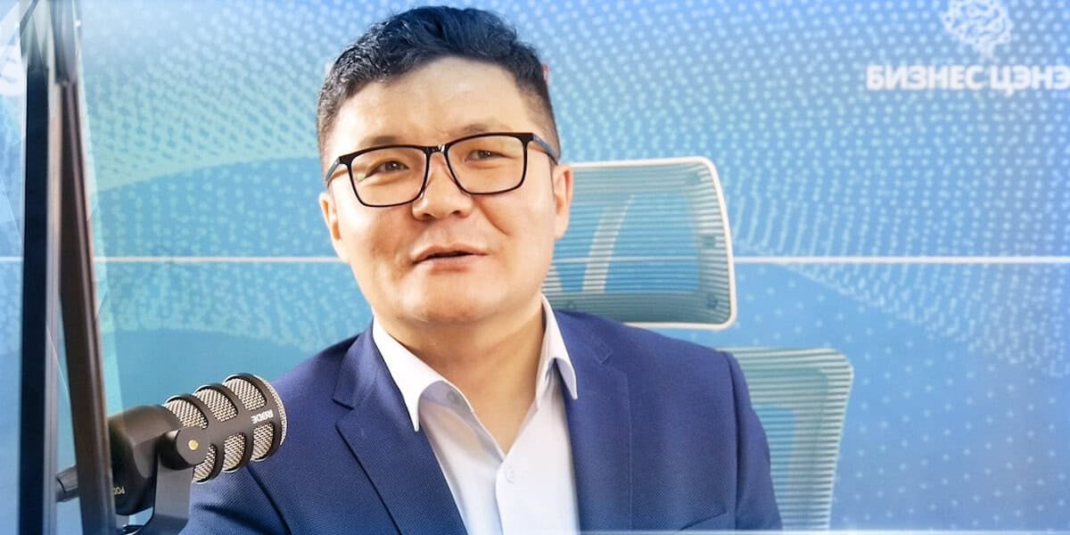 Нүдний шил, цэнхэр костюм өмссөн хүн микрофонд ярьж байна. Тэрээр хээ, бичвэр бүхий цэнхэр дэвсгэрийн өмнө сууж байгаа нь мэргэжлийн орчинд байгаа бололтой. - Business.MN