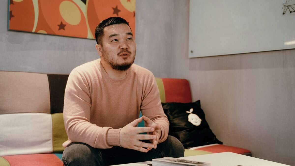 Богино хар үстэй, сахалтай, цайвар ягаан ноосон цамц өмссөн эрэгтэй өнгөт битүү буйдан дээр сууж байна. Тэрээр гараа атгаж, бизнесийнхээ талаар халуун сэтгэлээр ярьж байна. Арын дэвсгэр дээр цагаан самбар, ханан дээр өнгөлөг хийсвэр зураг байдаг. - Business.MN