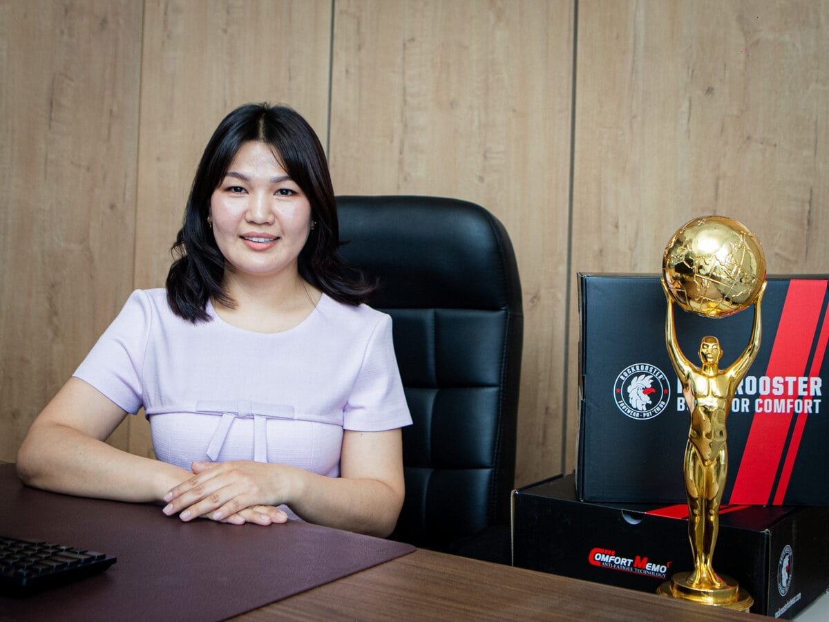 A woman in a light purple dress sits at an office desk, smiling. Beside her is a gold trophy shaped like a person holding up a globe, with black and red boxes stacked nearby. The background is a wooden wall.
